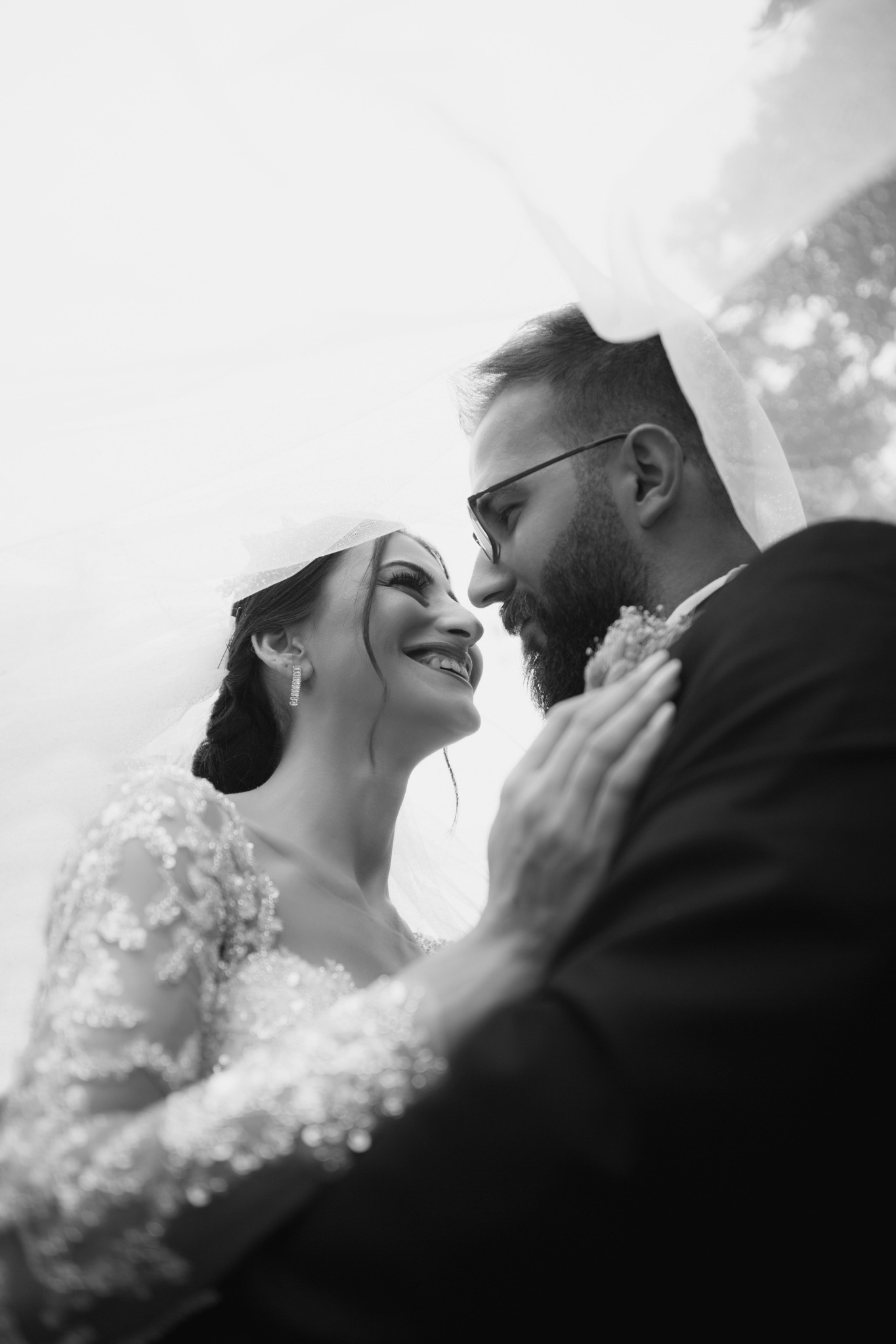 Black-and-white portrait of a bride and groom smiling at each other beneath a veil